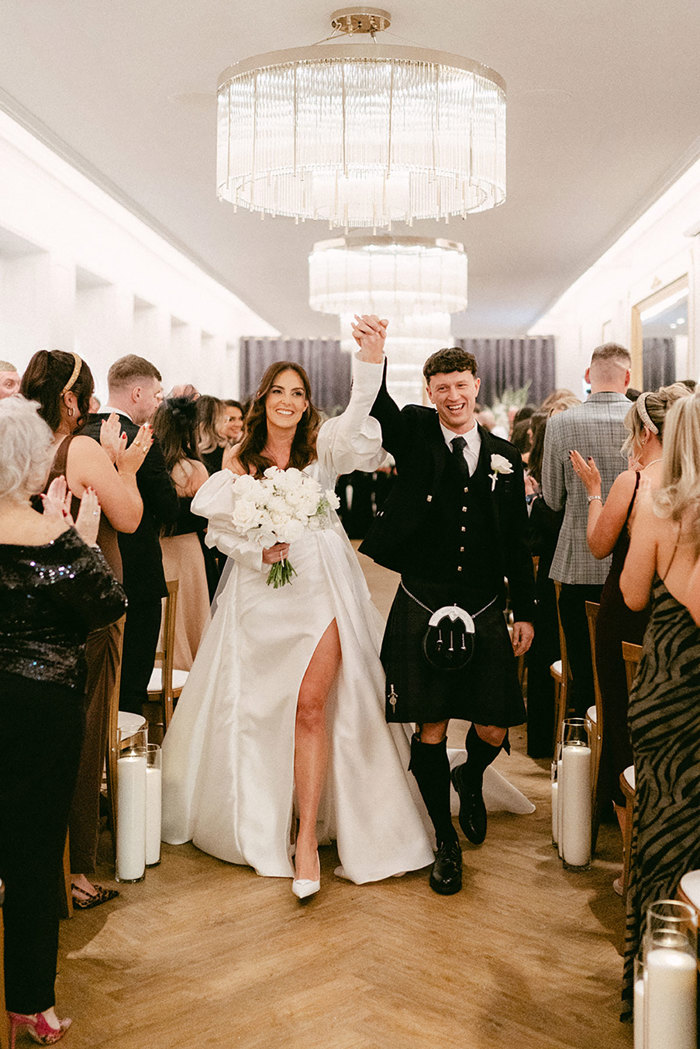 brunette bride in high leg slit wedding dress with puff sleeves and groom in full dark tartan kilt outfit walk up the aisle of a wedding ceremony with their hands intertwined above their heads