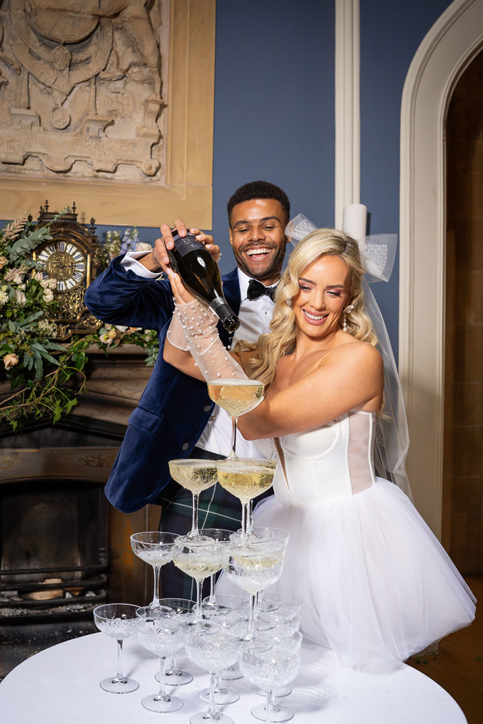 A groom wearing a navy suit jacket and a bride wearing a short wedding dress with tulle gloves and a bow pour prosecco into a tower of glasses