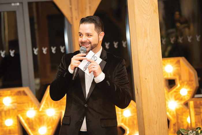 Man wearing black suit jacket with microphone in front of large letters with lights