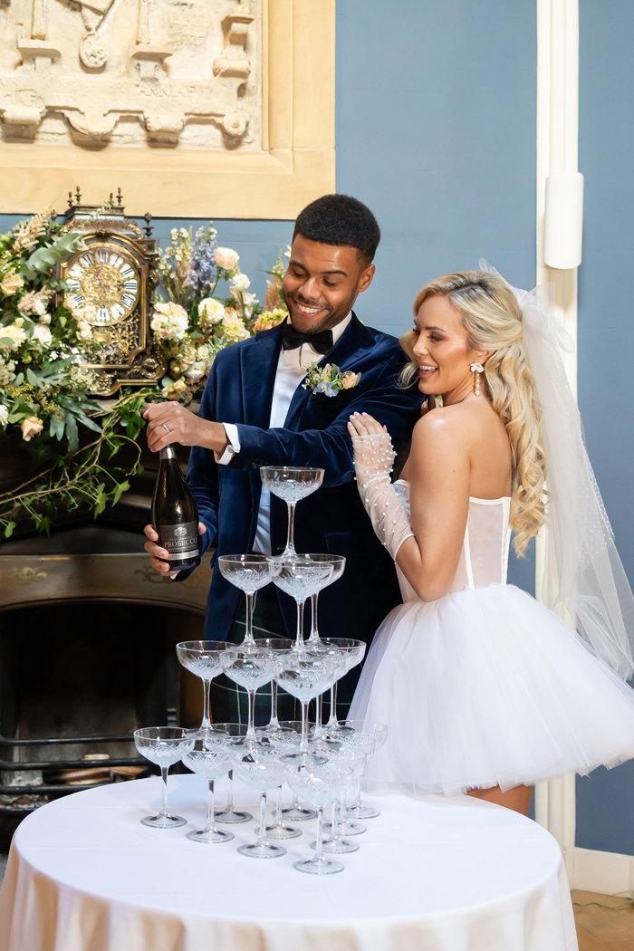 A groom wearing a navy suit jacket and a bride wearing a short wedding dress with tulle gloves and a bow pour prosecco into a tower of glasses