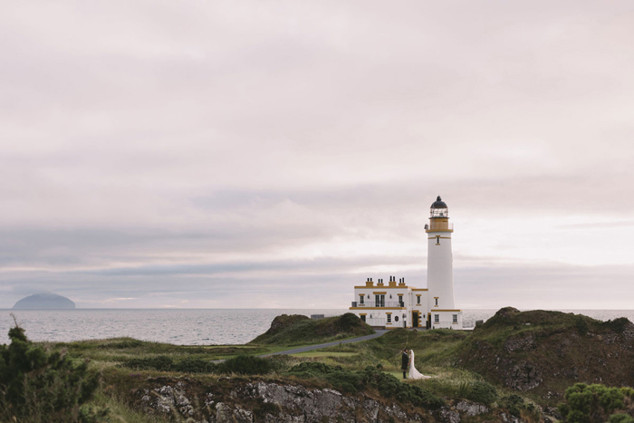 Bride and groom stand with a dramatic backdrop and lighthouse in the background