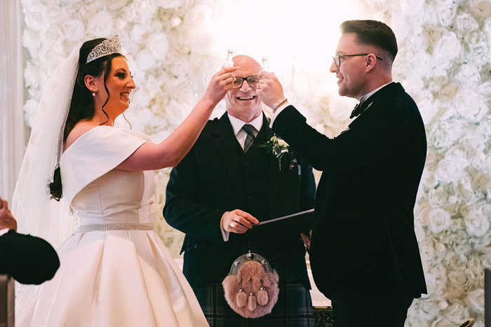 A person wearing a wedding dress and a person in a suit toasting with small glass bottles. A man in a kilt stands behind them.