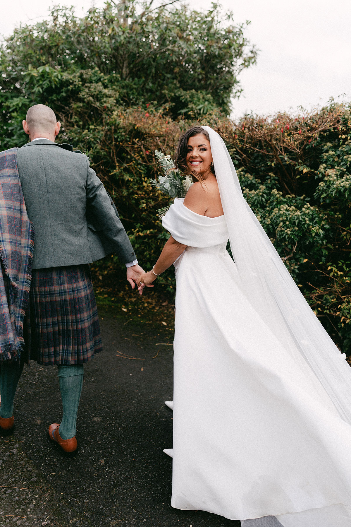 bride with long veil and off the shoulder wedding dress holds hands with groom in green, blue and purple tartan kilt outfit with green jacket, looking back over her shoulder and smiling