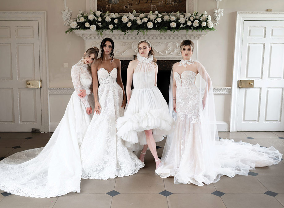 four women in bridalwear stand alongside each other in a horizontal line striking various poses to show off their dresses in front of an unused white marble fireplace decorated with white roses