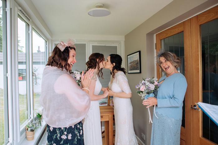 Two Brides Kissing As Their Mothers Clap