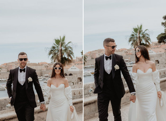 a bride and groom wearing formal attire and sunglasses walking in Dubrovnik with city skyline and boats in background