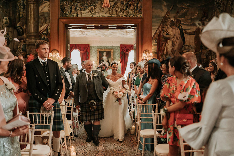 Wedding guests on either side of an aisle turn around to look at a bride walking in arm in arm with her father