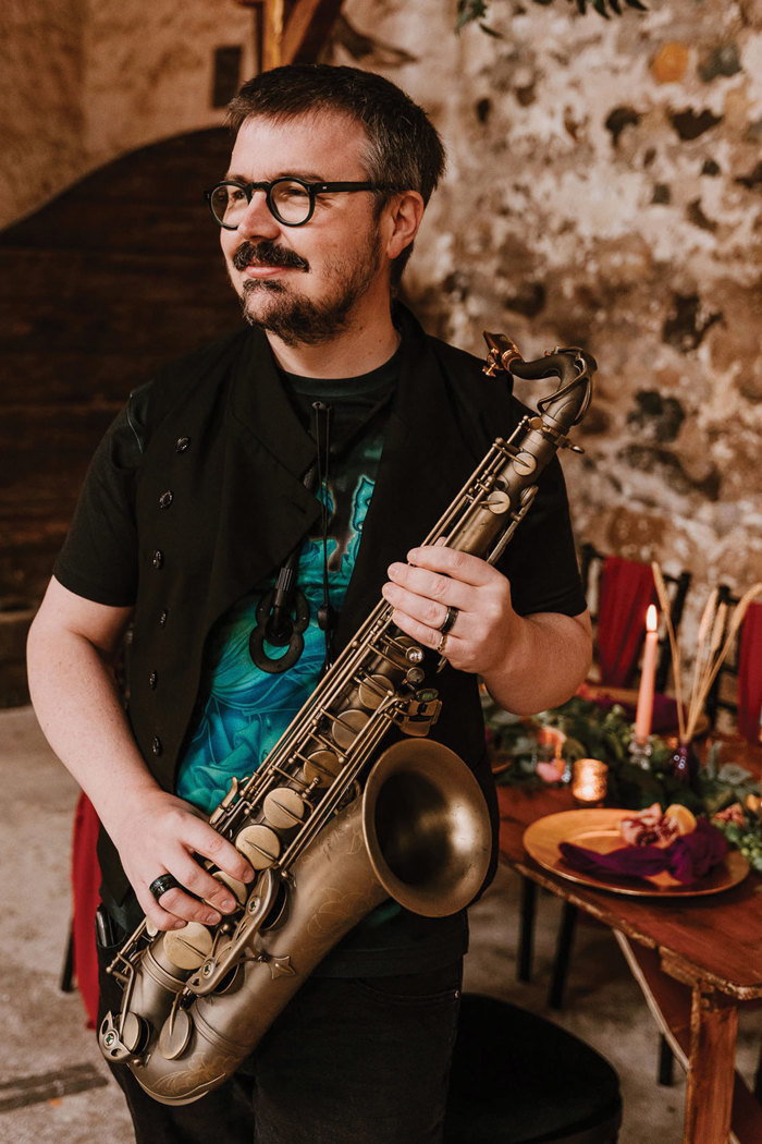 dark haired man with facial hair and glasses plays saxophone stood in a brick-walled room with a formal dining table in the back