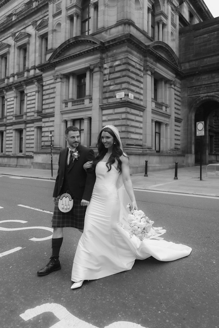 A black and white photo of a bride and groom walking arm in arm across a road