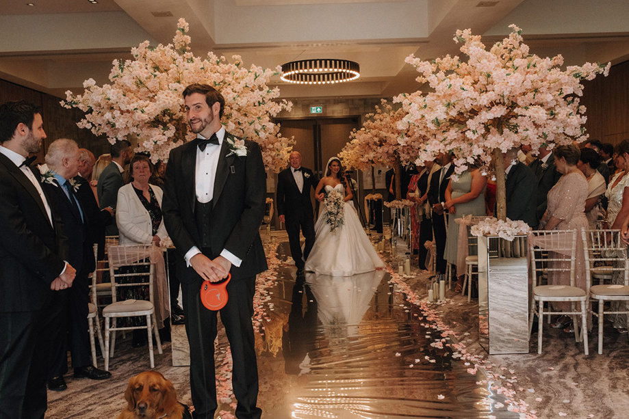 a groom at the top of the aisle with a dog on a lead as the bride walks towards him on the arm of a man in a suit. The aisle is decorated with cherry blossom trees either side.