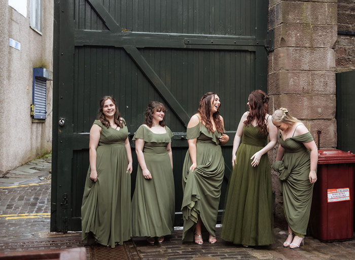 five bridesmaids wearing dark green dresses standing on a cobbled pavement against a large black wooden door