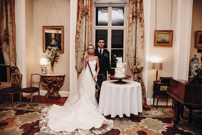  A Bride Holding A Sword Poses With A Groom Next To A Wedding Cake at Winton Castle