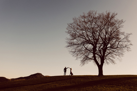 Groom spins his bride at sunset on a hill with a large bare tree beside them