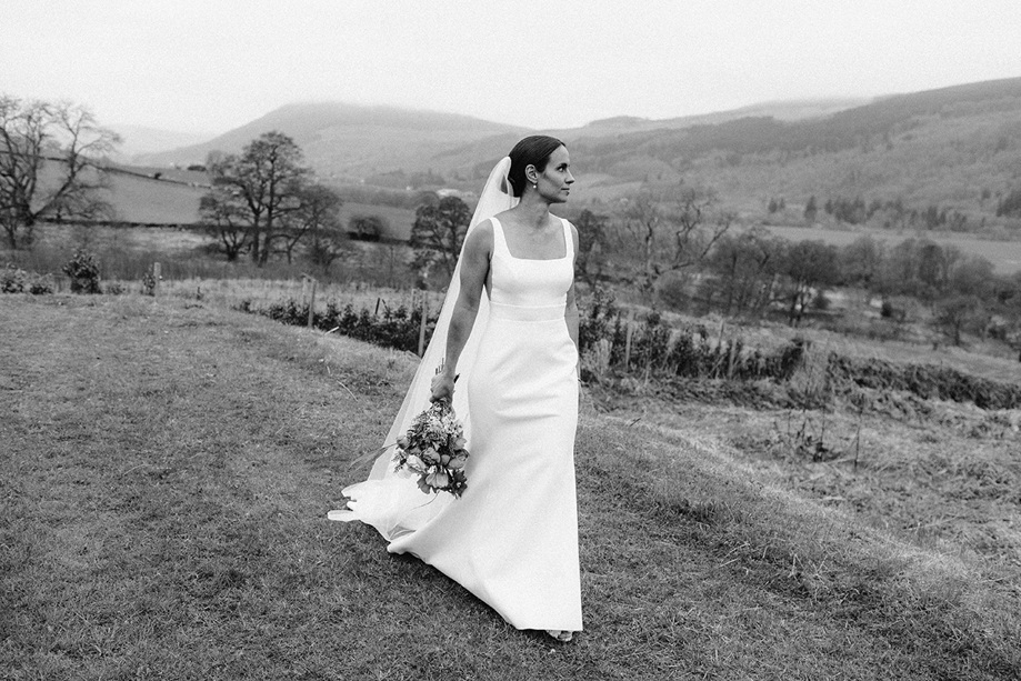 Bride walking across grass with bouquet in hand and hills in background