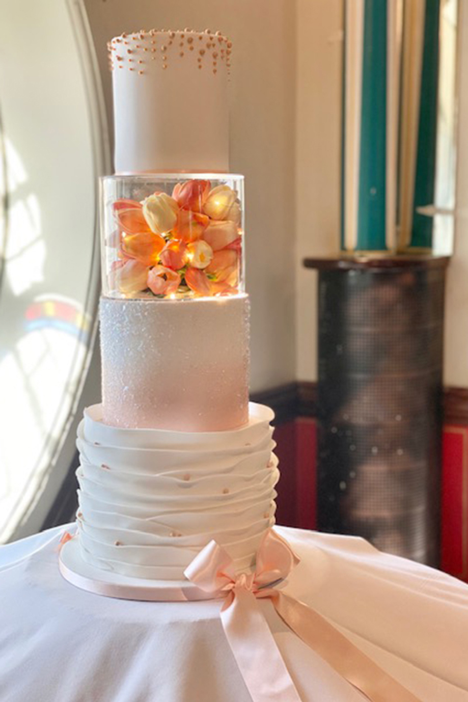 Pink and white four-tier cake with pink pearls and acrylic, tulip and fairy light-filled tier and pink bow around base