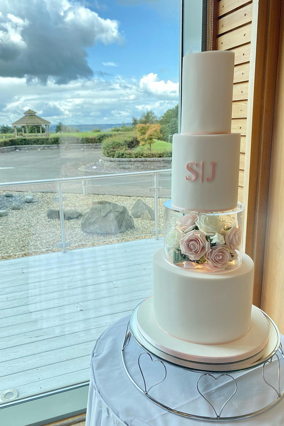 Pink and white four-tier cake at window with letters S and J on second tier, with acrylic tier filled with pink and white roses and fairy lights