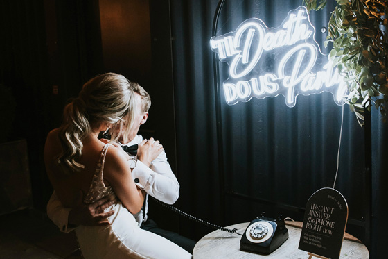 Bride and groom leave a message on their audio guest book with "Til Death Do Us Party" sign in background