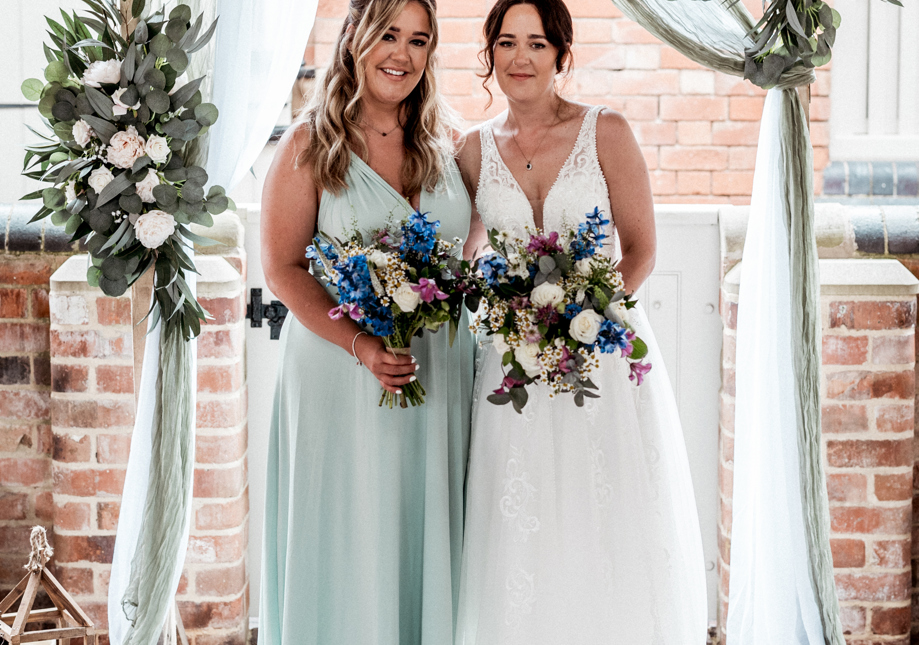 Bride and bridesmaid stand beneath a floral wedding arch with flowing white and sage drapes. The bride is in a white lace gown, and her bridesmaid is dressed in a pale green gown. Both hold lush bouquets featuring white roses, purple accents, and blue flowers, with a rustic brick background.”