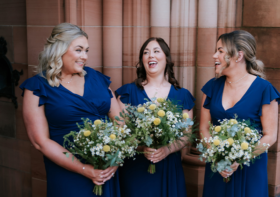 Bridesmaids in navy dresses laughing holding blue, yellow and white floral bouquets