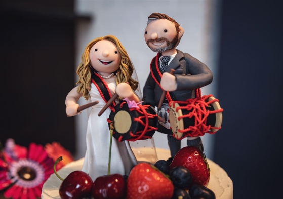 A polymer clay bride and groom playing taiko drums on top of a wedding cake surrounded by fruit