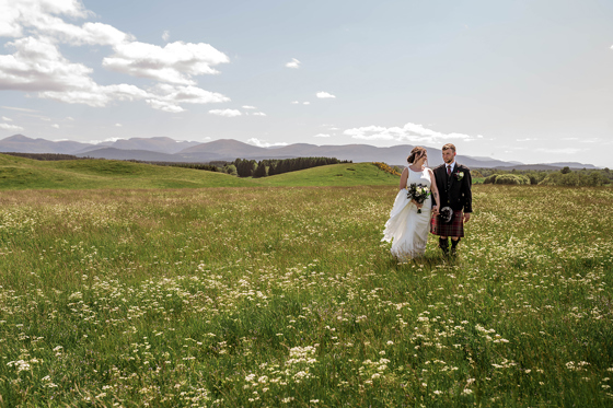Bride and groom walk together across grass with mountains in distance behind them
