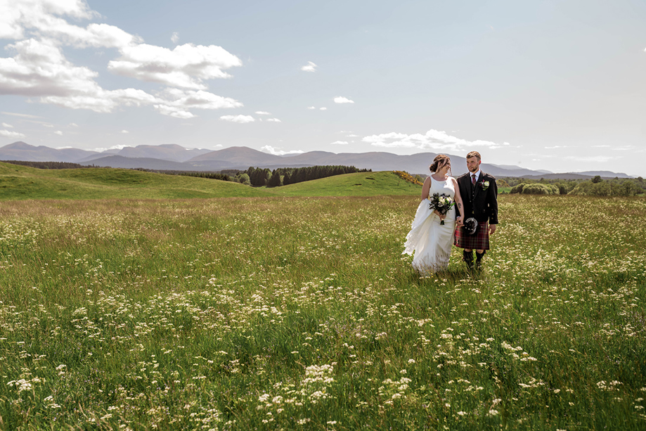 Bride and groom walk together across grass with mountains in distance behind them