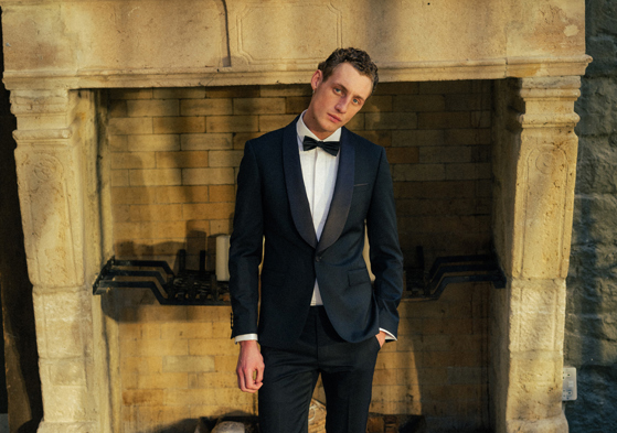 Man in black and white tuxedo in front of exposed stone fireplace