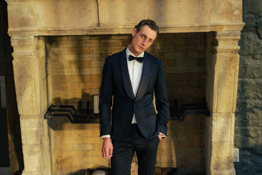 Man in black and white tuxedo in front of exposed stone fireplace