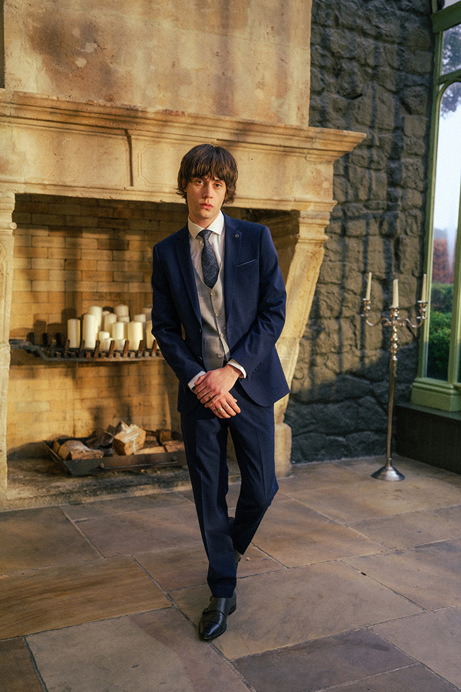 Man in three-piece grey and navy suit with tie in front of candle-filled exposed stonefireplace