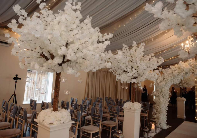 White floral trees on white pillars lining aisle, floral entrance archway with draped fabric ceiling covered in fairy lights