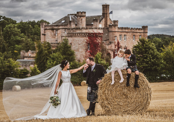 Groom kisses brides hand in field with two young children sitting atop hay bale with castle in background