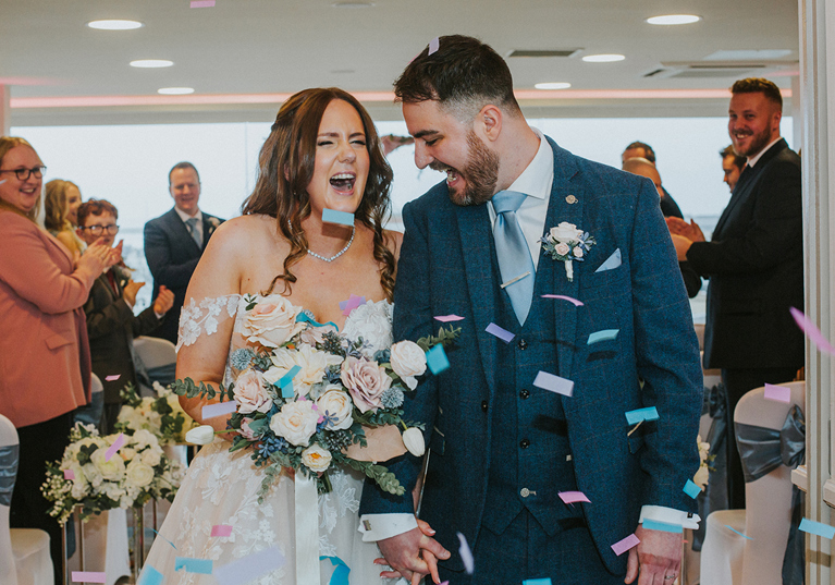 Bride and groom laugh as they walk through pastel coloured confetti thrown by guests