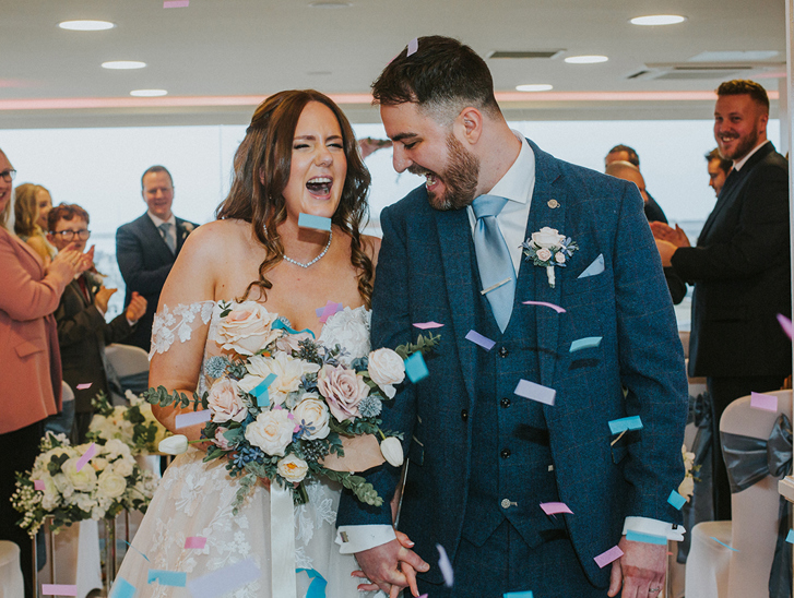 Bride and groom laugh as they walk through pastel coloured confetti thrown by guests