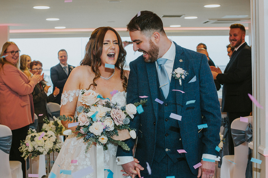 Bride and groom laugh as they walk through pastel coloured confetti thrown by guests
