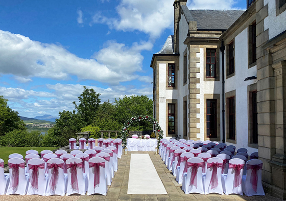 Outdoor ceremony set up with floral arch at altar and dark red organza bows on chairs