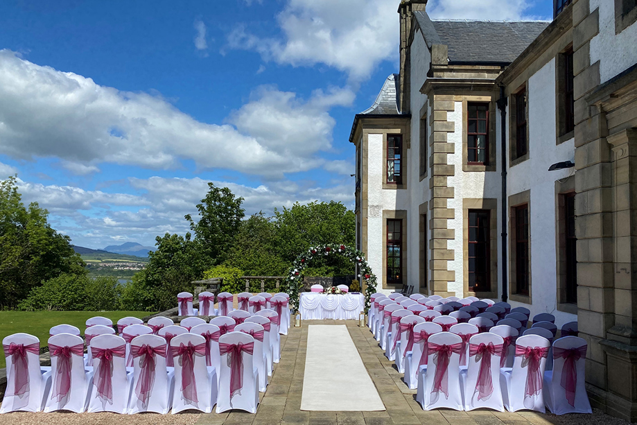 Outdoor ceremony set up with floral arch at altar and dark red organza bows on chairs