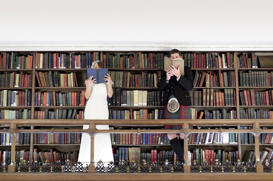 Bride and groom cover their faces with books whilst standing on the upper library