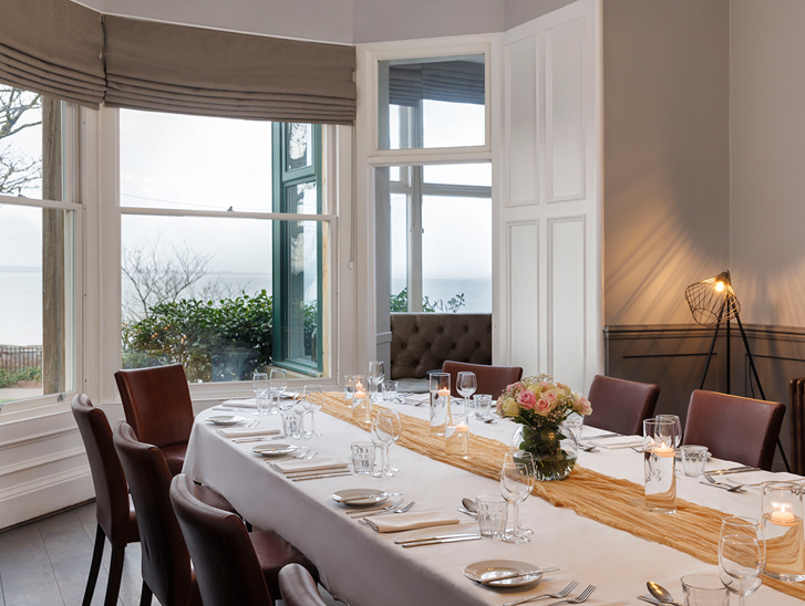 Wedding meal set up in the Macallan Room at Hotel du Vin St Andrews in Fife. Bay windows show the stunning views and the table is decorated with a mustard table runner and a small bouquet in the centre.