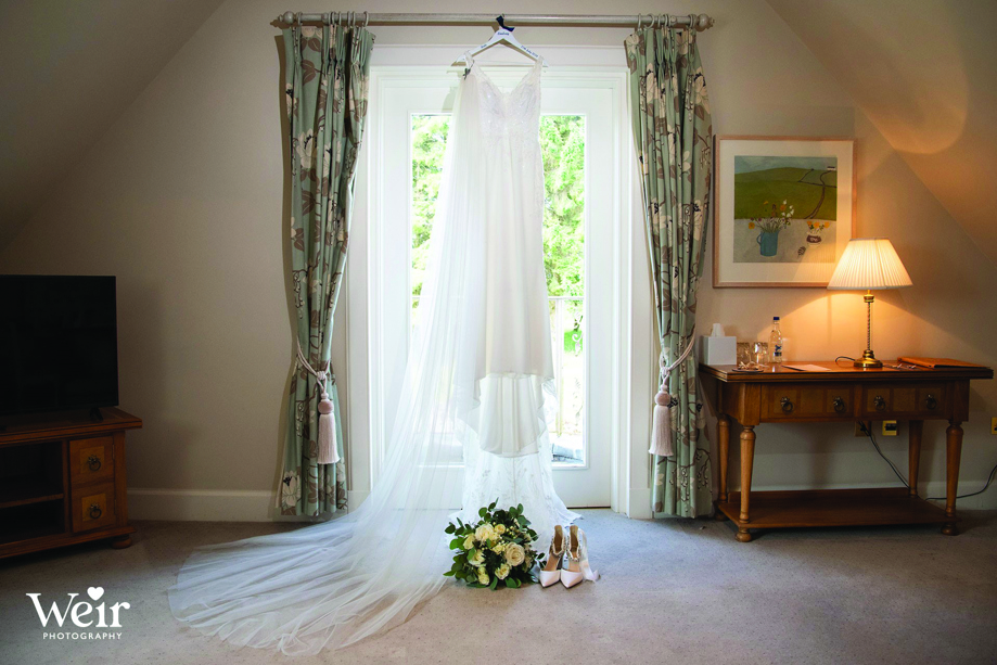 Image of wedding dress hanging at window with bouquet and bridal heels on floor by Weir Photography