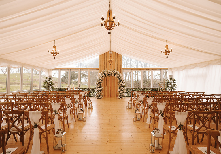 Wedding ceremony set up in the Garden Marquee at House of Elrick with golden chandeliers, chiavari chairs, silver lanterns and a flower arch.
