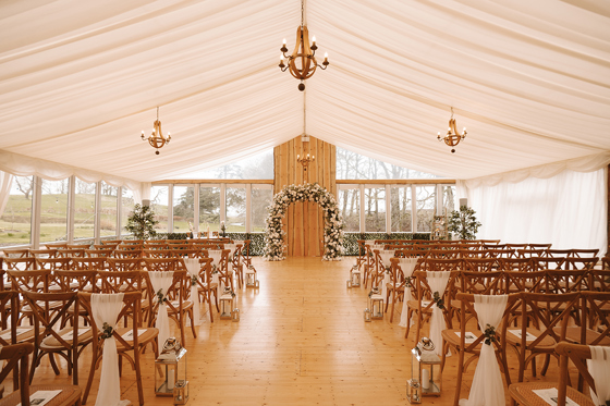 Wedding ceremony set up in the Garden Marquee at House of Elrick with golden chandeliers, chiavari chairs, silver lanterns and a flower arch.