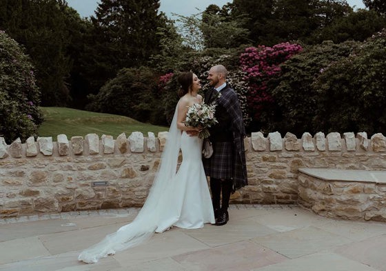 Bride and groom smile at one another in front of stone wall with leafy background