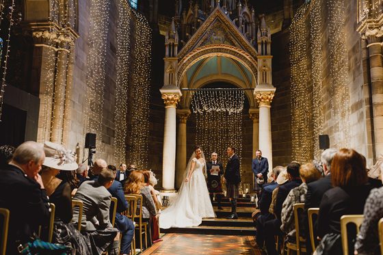 Bride smiles at guests at the altar with her husband-to-be and fairy lights decorating room