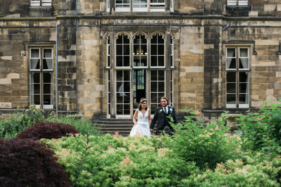 Bride and groom leaving building into gardens smiling and holding hands 