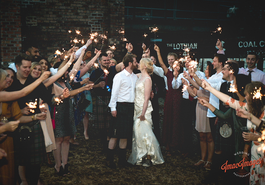 Bride and groom kiss with guests holding sparklers in background