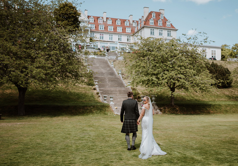 Bride looks back with sunglasses on walking with her groom towards Peebles Hydro Hotel