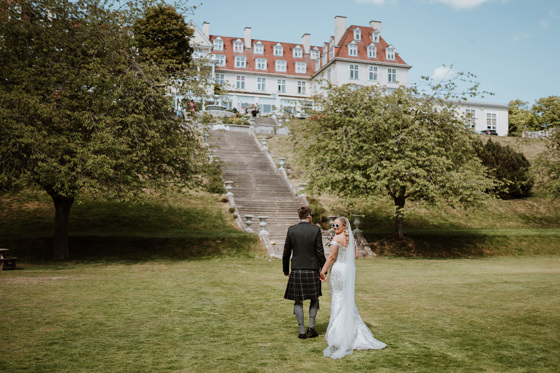 Bride looks back with sunglasses on walking with her groom towards Peebles Hydro Hotel