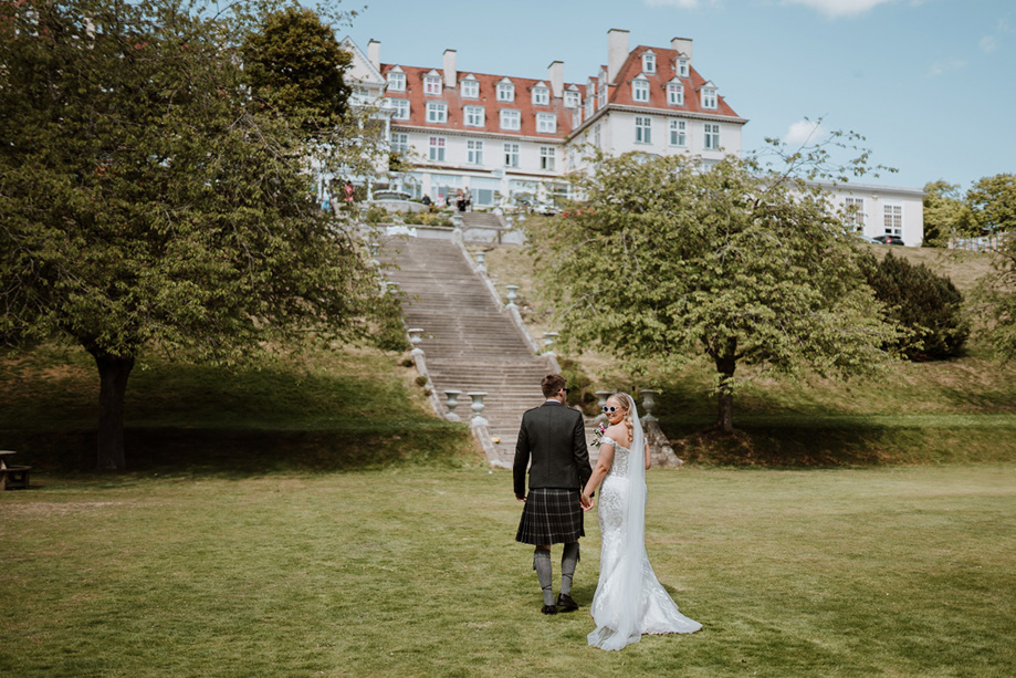 Bride looks back with sunglasses on walking with her groom towards Peebles Hydro Hotel