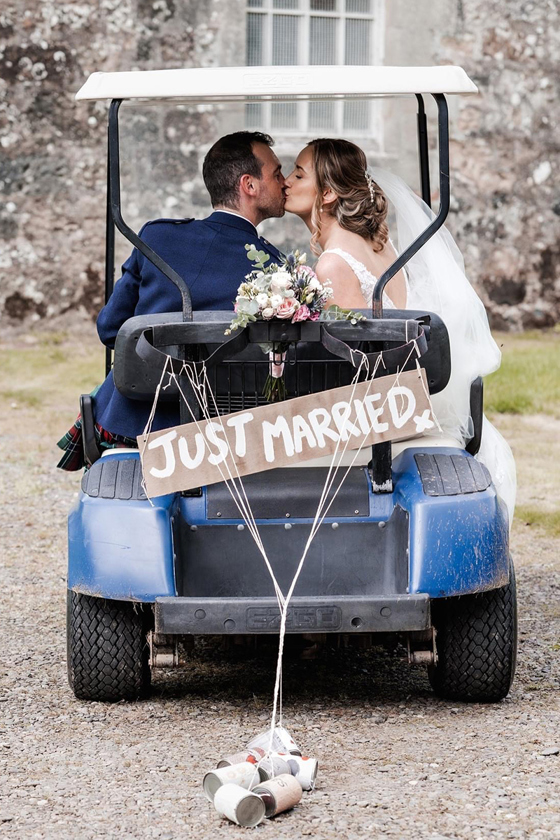 Bride and groom kiss as they sit in golf cart with "Just Married" sign and cans tied to back.