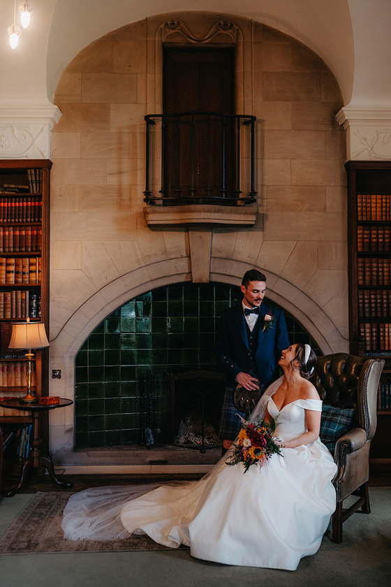 Bride sits on chair with bouquet smiling at her groom behind her in The Library at Rowallan Castle.